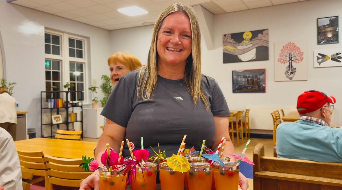 A smiling woman holding a tray of colourful drinks in a café-style room, with people sitting at tables behind her