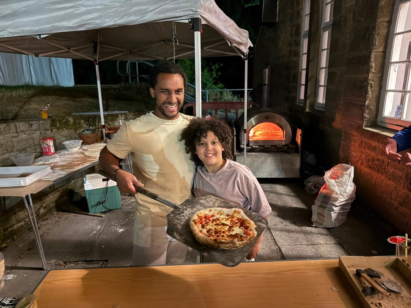 A smiling man and a child holding a freshly baked pizza beside the outdoor pizza oven